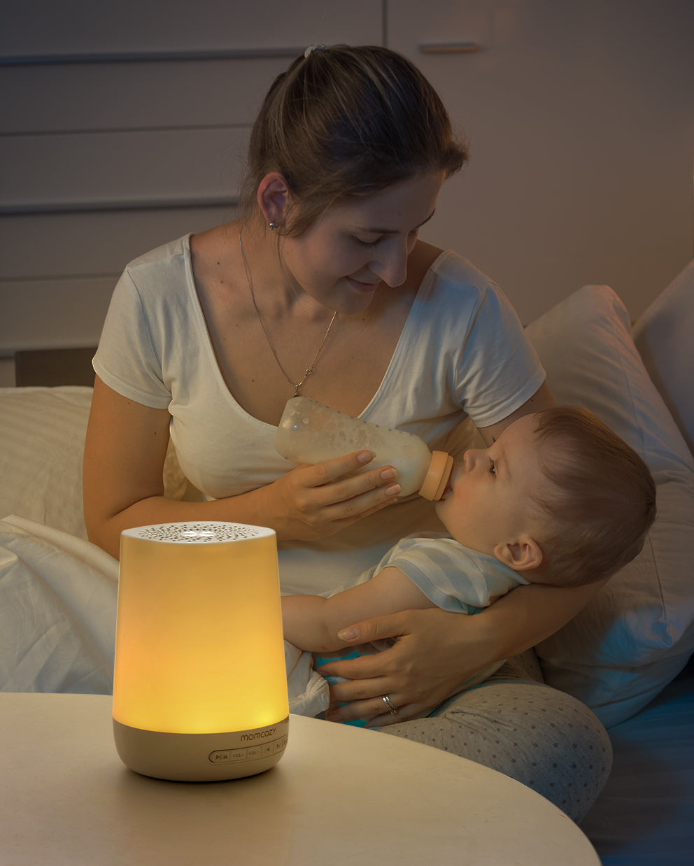 Mom feeding baby from bottle next to momcozy smart sound machine emitting warm yellow light