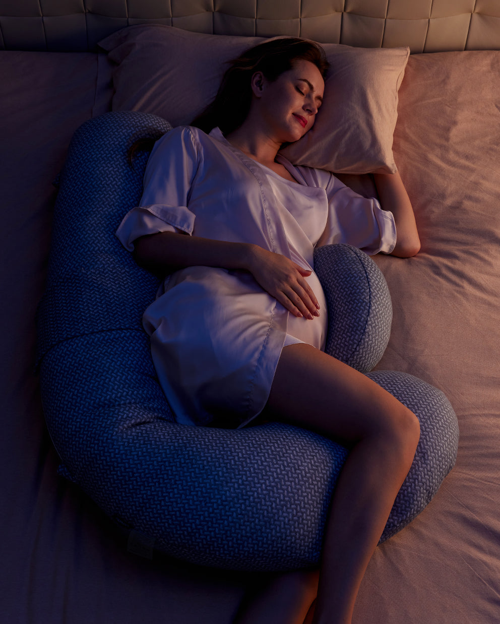Pregnant woman side-sleeping on gray Momcozy F-shaped pregnancy pillow for full body support and comfortable sleep.